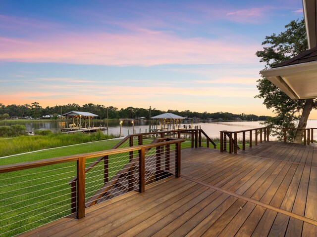 deck at dusk with a boat dock, a lawn, and a water view