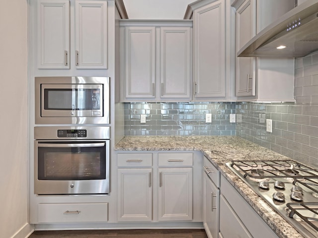 kitchen with white cabinets, wall chimney range hood, tasteful backsplash, light stone counters, and stainless steel appliances