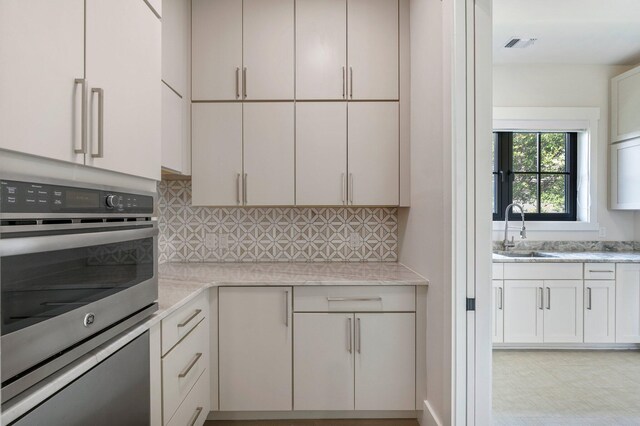 kitchen with oven, sink, tasteful backsplash, light stone counters, and white cabinetry