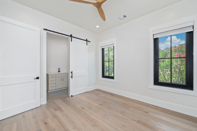 unfurnished bedroom with sink, ensuite bath, light hardwood / wood-style flooring, ceiling fan, and a barn door