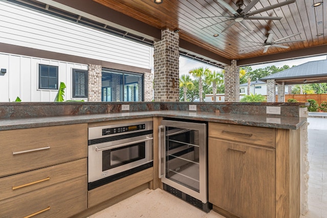 kitchen with ceiling fan, stainless steel oven, beverage cooler, dark stone countertops, and wood ceiling