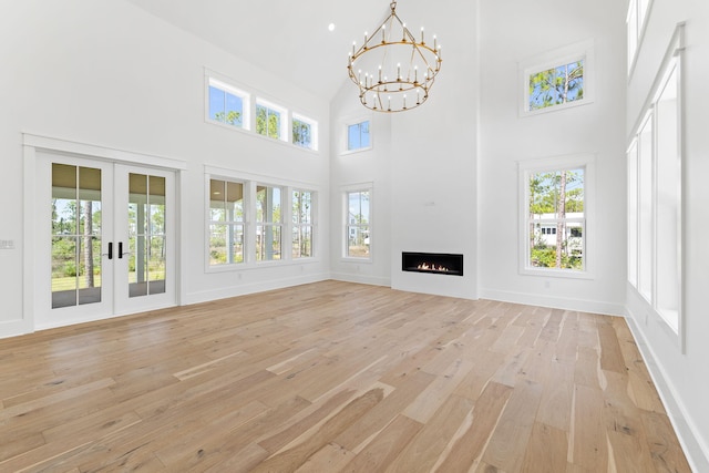 unfurnished living room featuring light wood finished floors, a towering ceiling, an inviting chandelier, a lit fireplace, and french doors