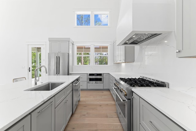 kitchen with high end appliances, custom range hood, light stone counters, gray cabinets, and a sink