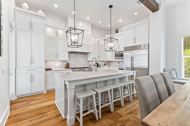 kitchen with pendant lighting, white cabinets, built in appliances, light stone countertops, and a center island with sink