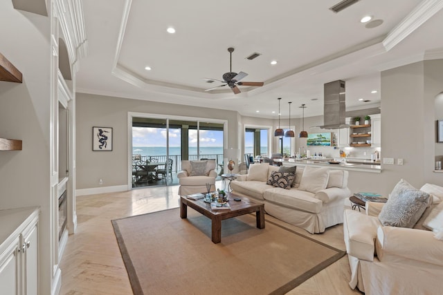 living room with ceiling fan, a tray ceiling, light parquet floors, ornamental molding, and a water view