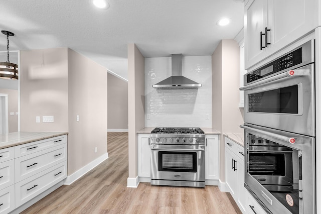 kitchen featuring wall chimney exhaust hood, stainless steel appliances, light stone counters, pendant lighting, and white cabinets