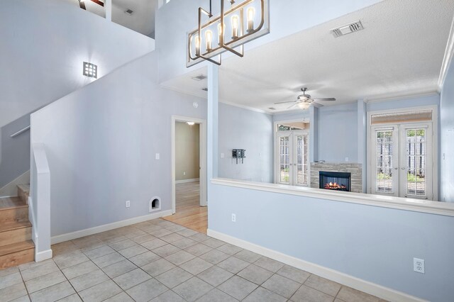 interior space featuring light tile patterned flooring, ceiling fan with notable chandelier, crown molding, french doors, and a stone fireplace