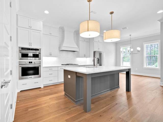 kitchen with custom range hood, hanging light fixtures, light hardwood / wood-style floors, white cabinets, and a center island with sink