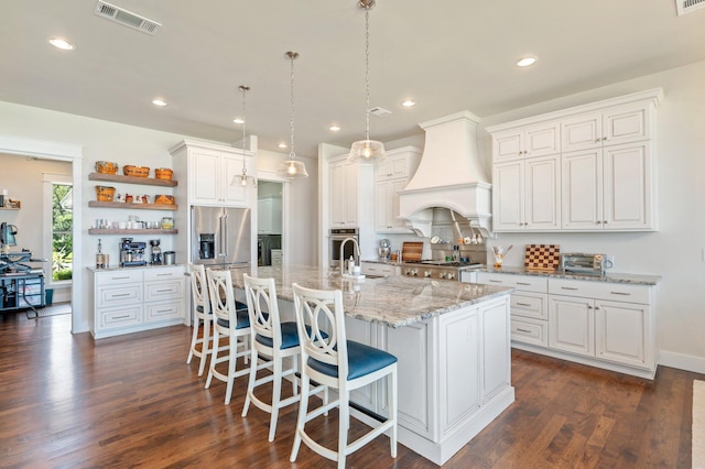 kitchen featuring a center island with sink, visible vents, custom range hood, hanging light fixtures, and white cabinetry