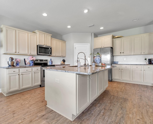 kitchen with light wood-type flooring, light stone counters, a center island with sink, and appliances with stainless steel finishes