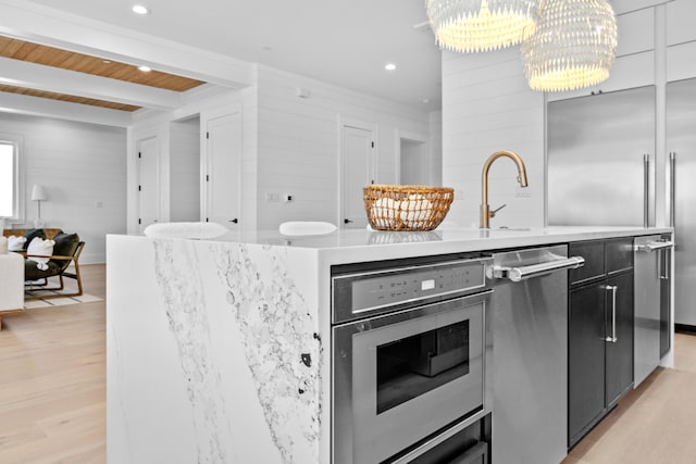 kitchen featuring an inviting chandelier, a center island with sink, light wood-type flooring, appliances with stainless steel finishes, and beamed ceiling