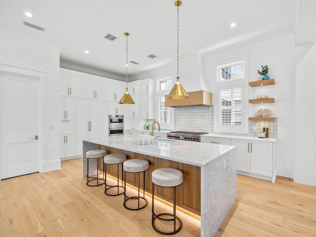 kitchen featuring light stone countertops, custom range hood, a kitchen island with sink, light hardwood / wood-style flooring, and white cabinetry