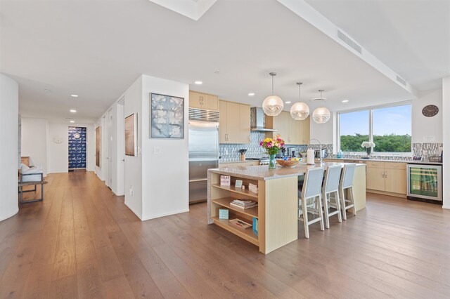 kitchen with an island with sink, wall chimney exhaust hood, stainless steel built in refrigerator, wine cooler, and backsplash