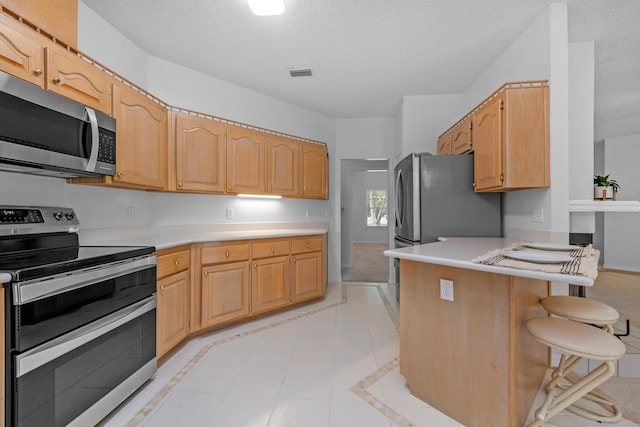 kitchen featuring appliances with stainless steel finishes, a kitchen breakfast bar, a textured ceiling, light brown cabinetry, and kitchen peninsula