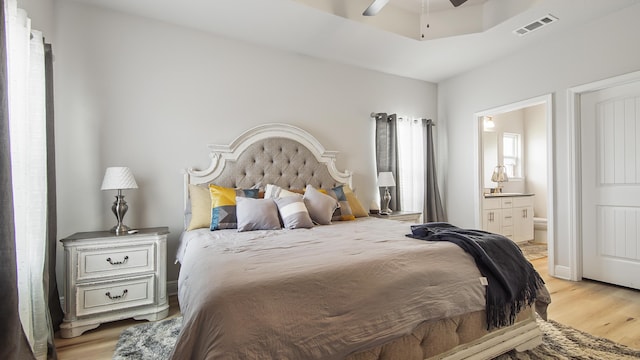 bedroom featuring a raised ceiling, visible vents, ceiling fan, ensuite bath, and light wood-type flooring