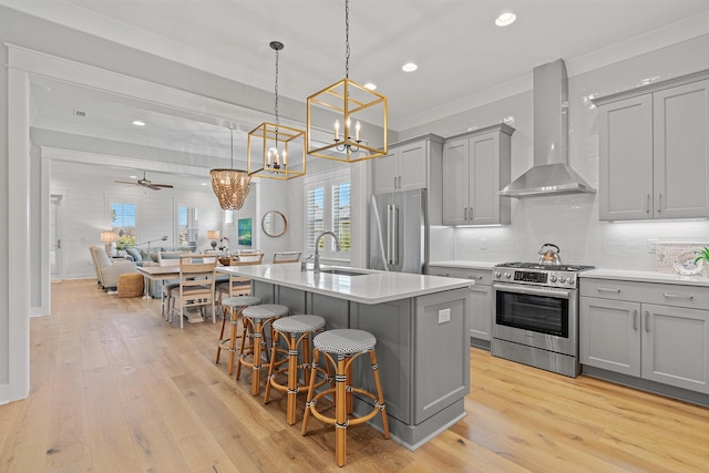 kitchen with appliances with stainless steel finishes, ceiling fan with notable chandelier, wall chimney range hood, pendant lighting, and an island with sink