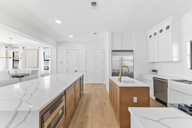 kitchen featuring built in appliances, pendant lighting, light stone countertops, a center island with sink, and light hardwood / wood-style floors