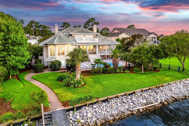 back house at dusk with a water view, a balcony, a fenced in pool, and a lawn