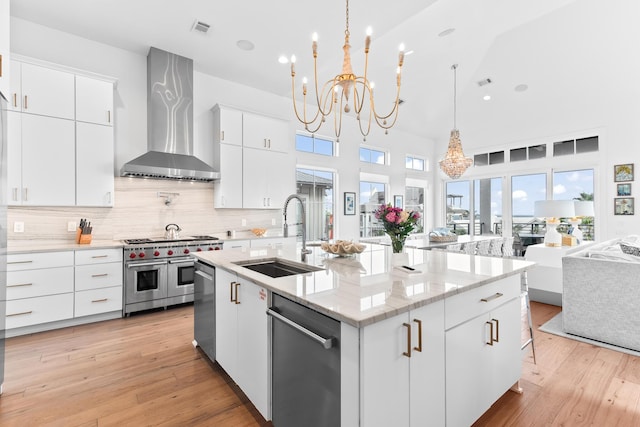 kitchen with wall chimney range hood, sink, a kitchen island with sink, appliances with stainless steel finishes, and white cabinets