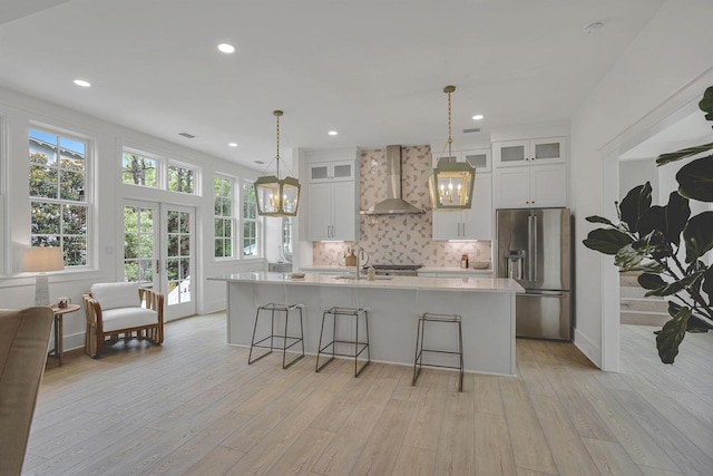 kitchen featuring stainless steel refrigerator with ice dispenser, wall chimney exhaust hood, a center island with sink, and light hardwood / wood-style floors