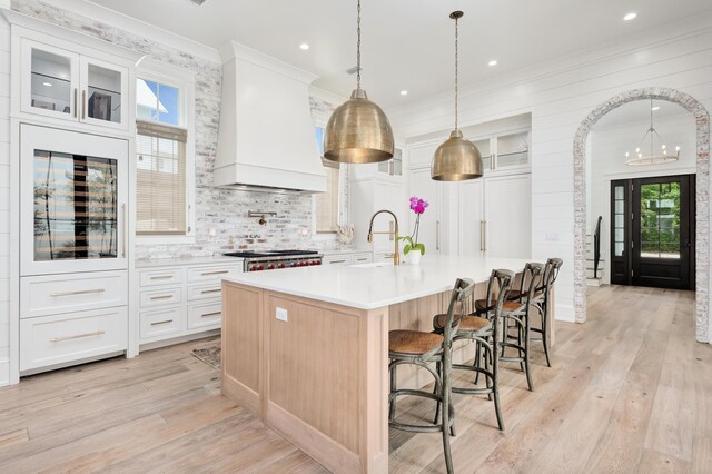 kitchen with custom range hood, a center island with sink, white cabinetry, and a wealth of natural light