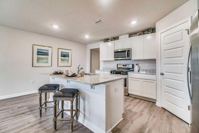 kitchen with light stone counters, a textured ceiling, stainless steel appliances, a center island with sink, and white cabinets