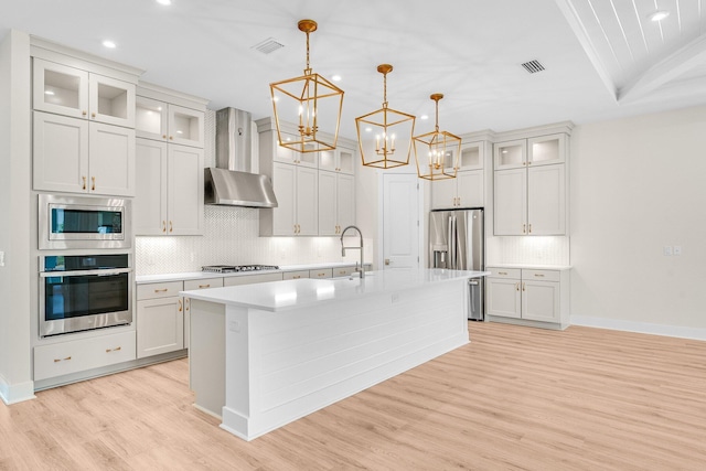 kitchen with glass insert cabinets, a kitchen island with sink, stainless steel appliances, light countertops, and wall chimney range hood