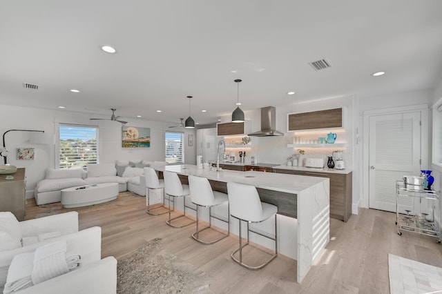 kitchen with wall chimney exhaust hood, a breakfast bar, decorative light fixtures, a kitchen island with sink, and light hardwood / wood-style floors