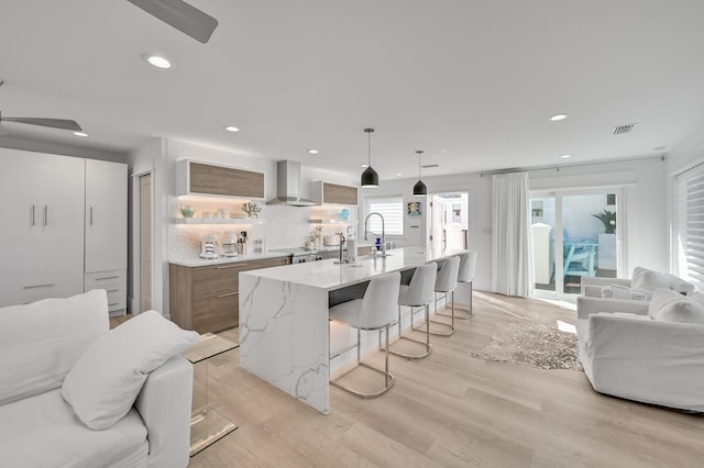 kitchen with decorative light fixtures, an island with sink, a kitchen breakfast bar, light wood-type flooring, and wall chimney exhaust hood