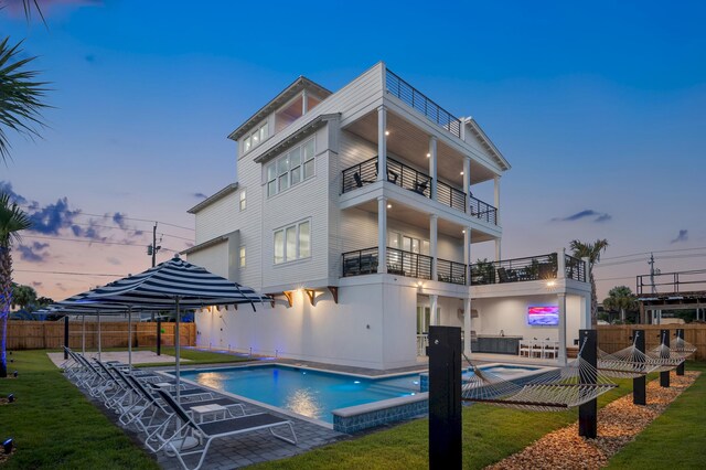 back house at dusk featuring a balcony, a patio area, a lawn, and a fenced in pool