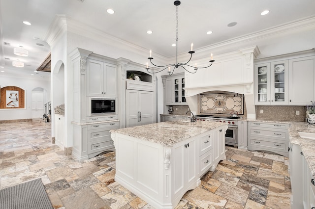 kitchen featuring a center island with sink, hanging light fixtures, light stone countertops, built in microwave, and tasteful backsplash