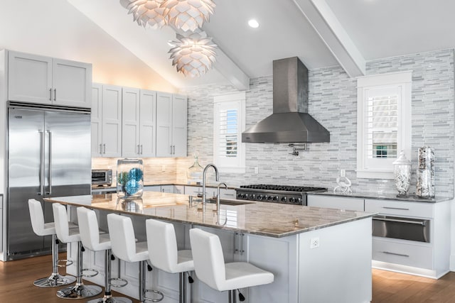 kitchen featuring an island with sink, sink, stainless steel built in fridge, light stone counters, and wall chimney range hood