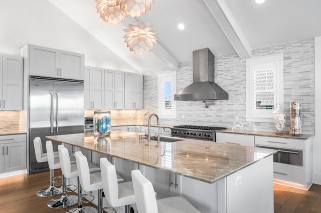 kitchen featuring high quality appliances, an island with sink, sink, light stone countertops, and wall chimney range hood