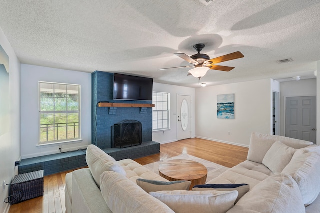 living room with light wood-type flooring, a textured ceiling, ceiling fan, a brick fireplace, and brick wall