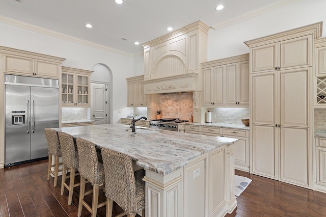 kitchen with light stone counters, arched walkways, custom exhaust hood, cream cabinets, and stainless steel built in fridge