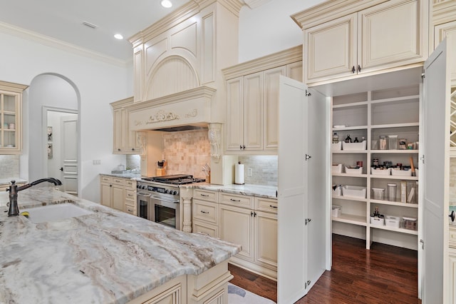 kitchen featuring arched walkways, range with two ovens, cream cabinets, a sink, and crown molding