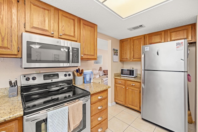 kitchen with light tile patterned floors, visible vents, brown cabinetry, light stone countertops, and stainless steel appliances