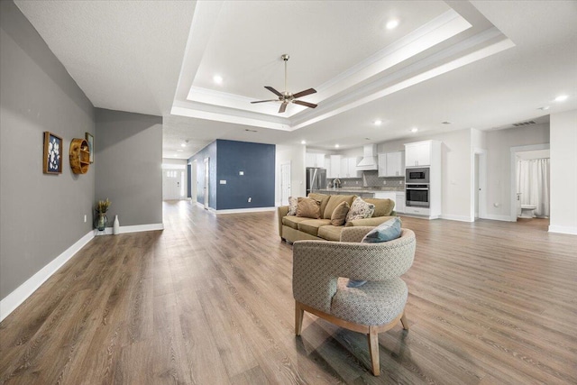 living room featuring ceiling fan, light wood-type flooring, ornamental molding, sink, and a raised ceiling