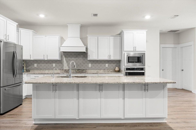 kitchen with stainless steel appliances, premium range hood, light hardwood / wood-style floors, decorative backsplash, and a kitchen island with sink