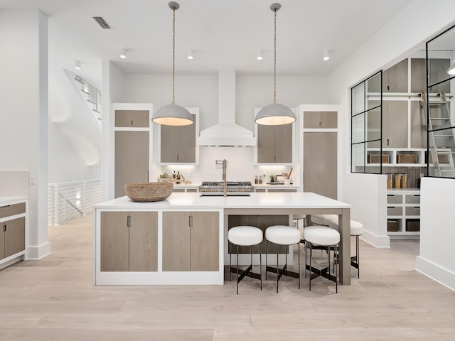 kitchen with gray cabinets, light wood-type flooring, custom exhaust hood, hanging light fixtures, and an island with sink