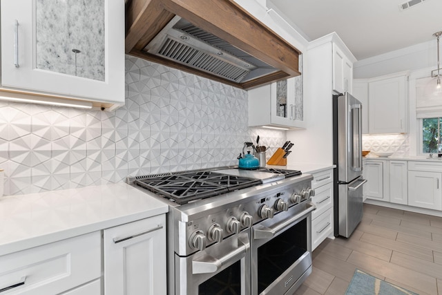 kitchen featuring backsplash, premium appliances, custom range hood, white cabinets, and decorative light fixtures