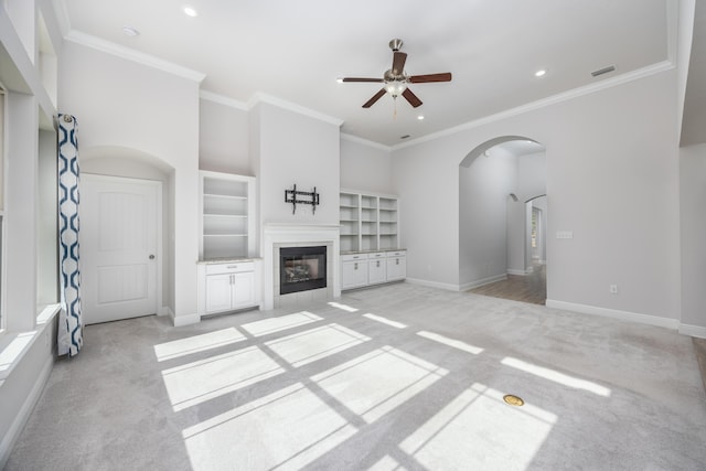 unfurnished living room featuring a tiled fireplace, ceiling fan, built in features, crown molding, and light colored carpet