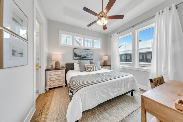 bedroom featuring multiple windows, light hardwood / wood-style flooring, ceiling fan, and a tray ceiling