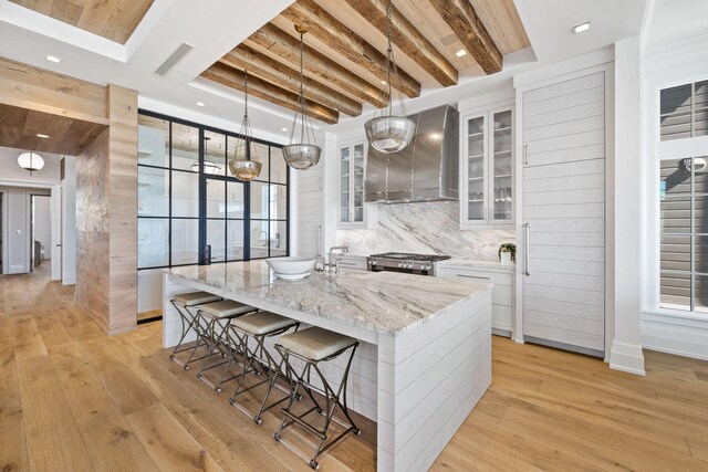 kitchen featuring a kitchen island with sink, wall chimney range hood, white cabinets, pendant lighting, and tasteful backsplash