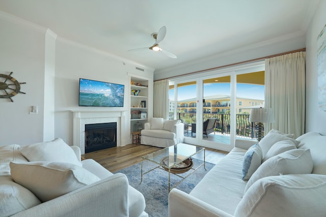 living room featuring light wood-type flooring, a fireplace, crown molding, built in features, and ceiling fan