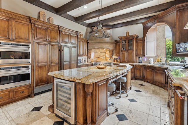 kitchen featuring light stone counters, an island with sink, beamed ceiling, pendant lighting, and beverage cooler