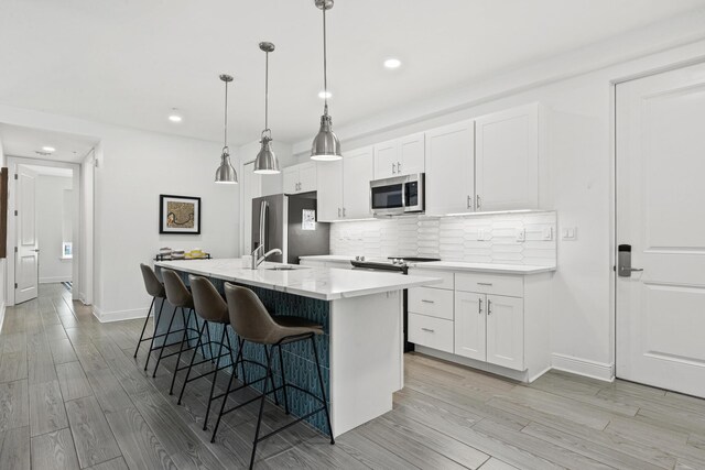 kitchen with appliances with stainless steel finishes, an island with sink, backsplash, white cabinetry, and light hardwood / wood-style flooring