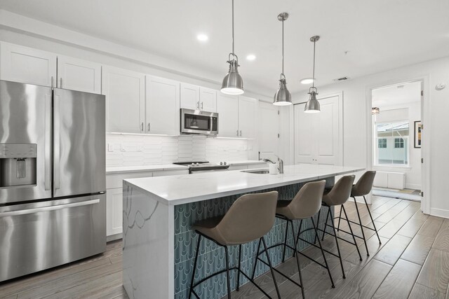 kitchen with appliances with stainless steel finishes, an island with sink, white cabinetry, backsplash, and wood-type flooring