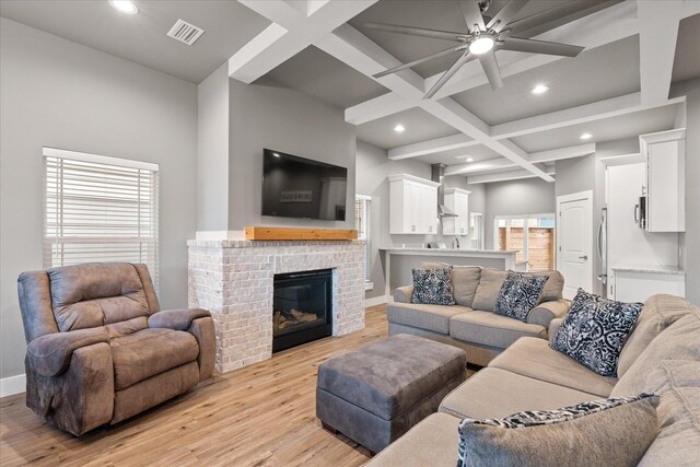 living room featuring a brick fireplace, light wood-type flooring, coffered ceiling, ceiling fan, and beam ceiling