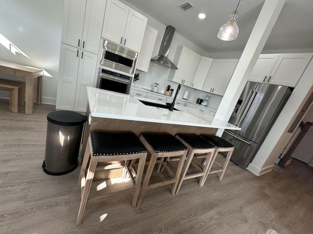 kitchen with wall chimney range hood, appliances with stainless steel finishes, a kitchen breakfast bar, white cabinets, and decorative light fixtures